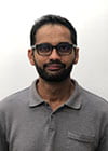 Harjit wears a moustache and beard, glasses, and a gray polo shirt.